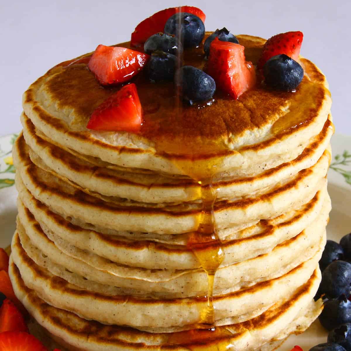 A tall stack of golden-brown eggless pancakes is topped with fresh blueberries and sliced strawberries. Maple syrup is being drizzled down the sides, creating a glossy sheen. The pancakes have a fluffy texture with slightly crisp edges. More berries are scattered around the plate, adding vibrant color.