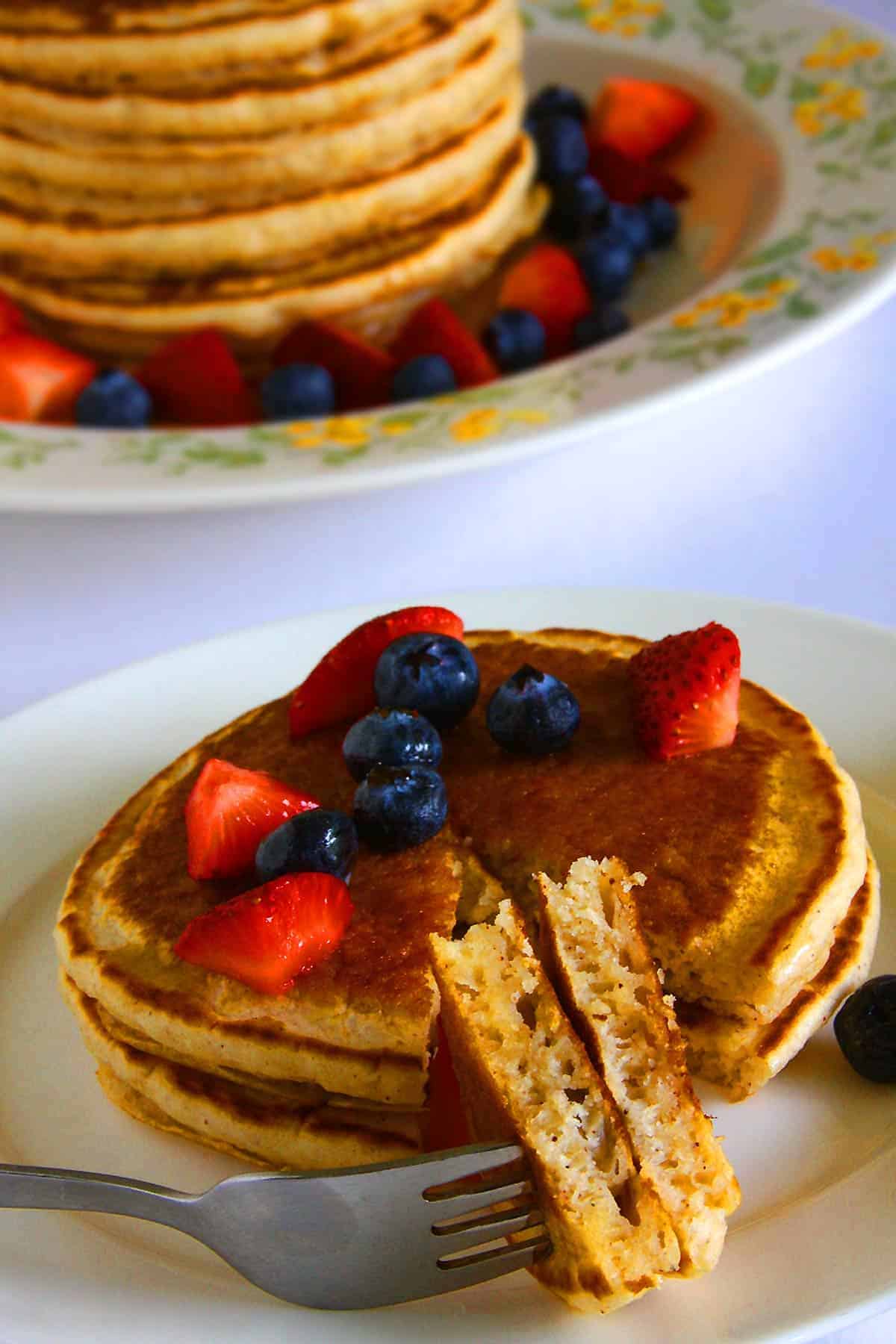 A stack of golden-brown eggless pancakes is served on a white plate, topped with fresh blueberries and sliced strawberries. A fork holds a bite-sized portion, revealing the pancakes' fluffy texture. The background is blurred with a white table setting.