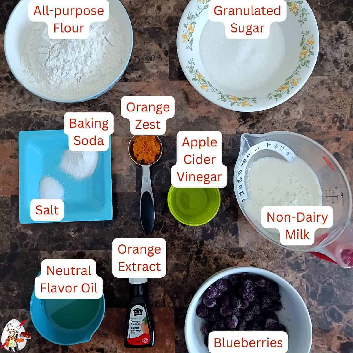 Ingredients for vegan blueberry muffins arranged on a marble surface, including flour, sugar, baking soda, salt, orange zest, vinegar, non-dairy milk, oil, extract, and blueberries.